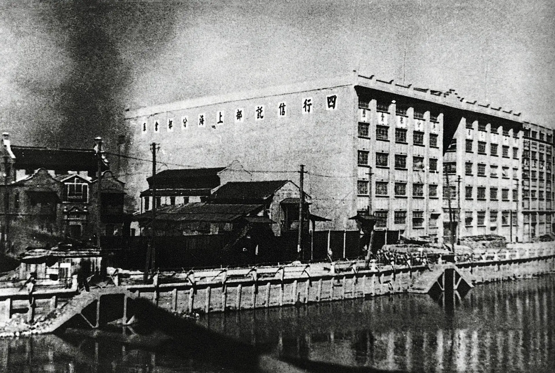 Sihang Warehouse across Suzhou Creek during the 1937 battle, with smoke rising beside the building.
