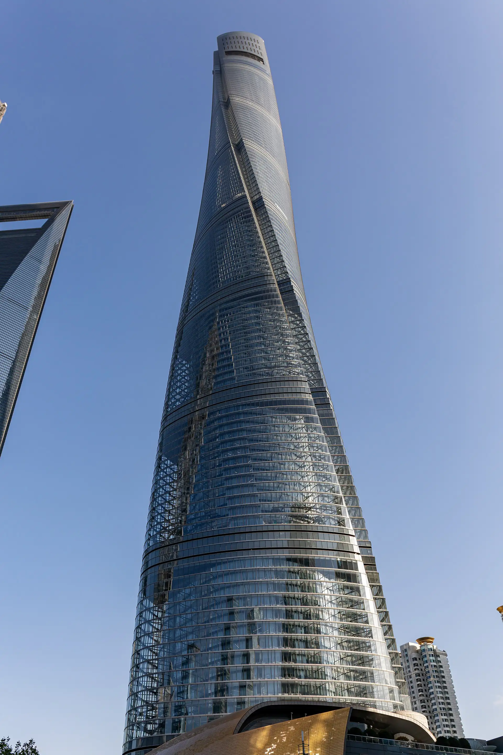 Shanghai Tower's twisting glass façade seen from below against a blue sky, with the World Financial Center at the left edge.