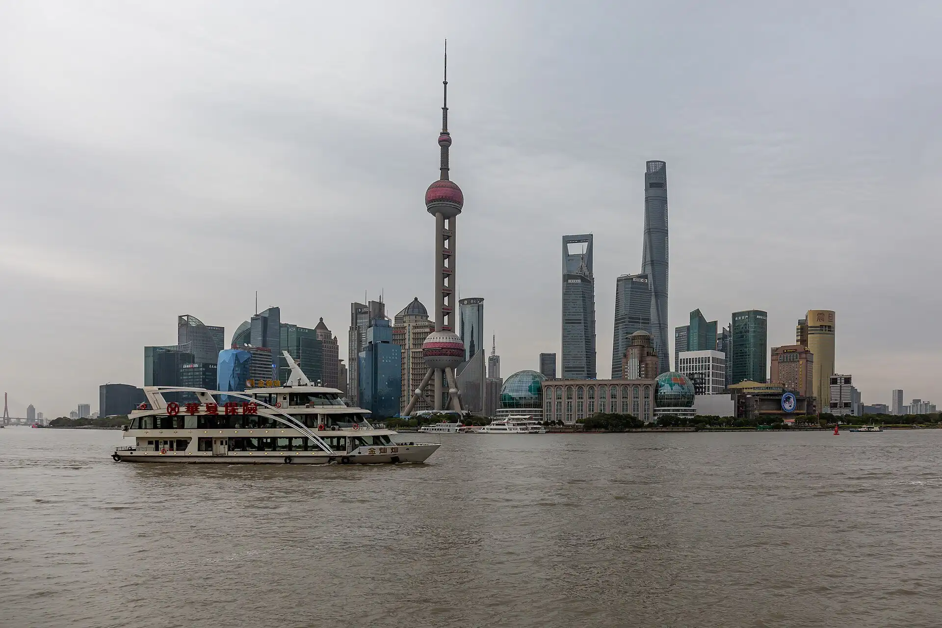 City skyline across a wide brown river under a grey sky, with a needle-shaped television tower with two pink spheres in front of three much taller skyscrapers, and a white tour boat on the water at left.