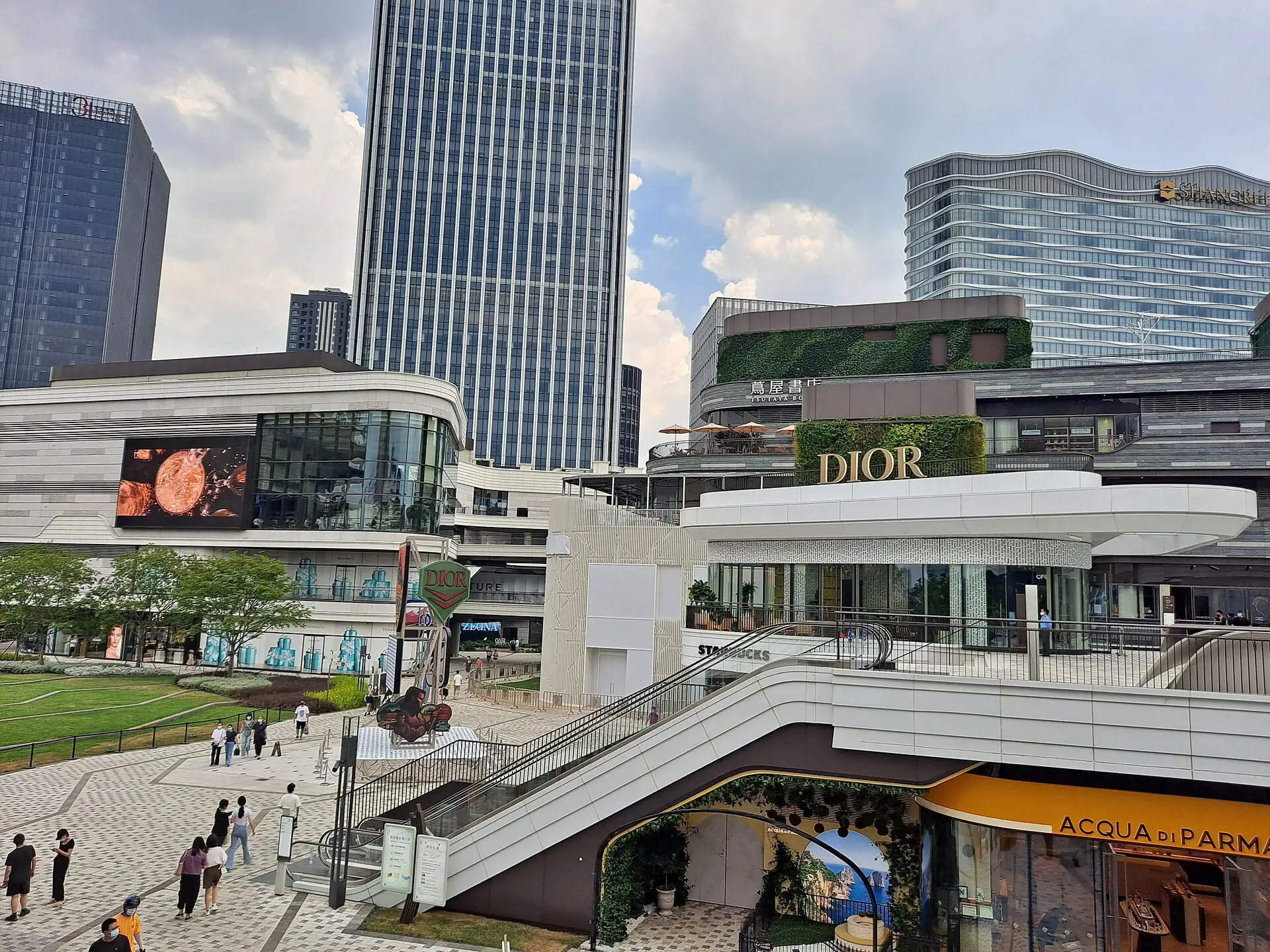 Open-air shopping centre with terraces, escalators and shop fronts, a lawn and people walking on a paved square, and tall office buildings behind.