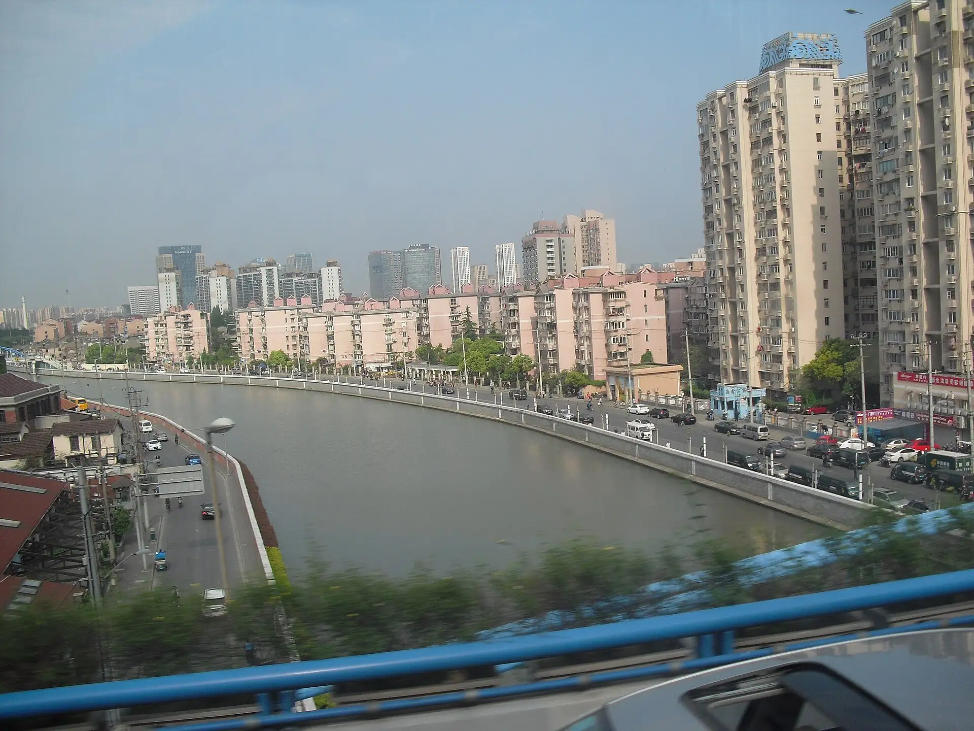 A curving river with concrete flood walls, a road along the far bank lined with pink apartment blocks and taller towers behind, seen from an elevated road with a blurred blue railing in the foreground.