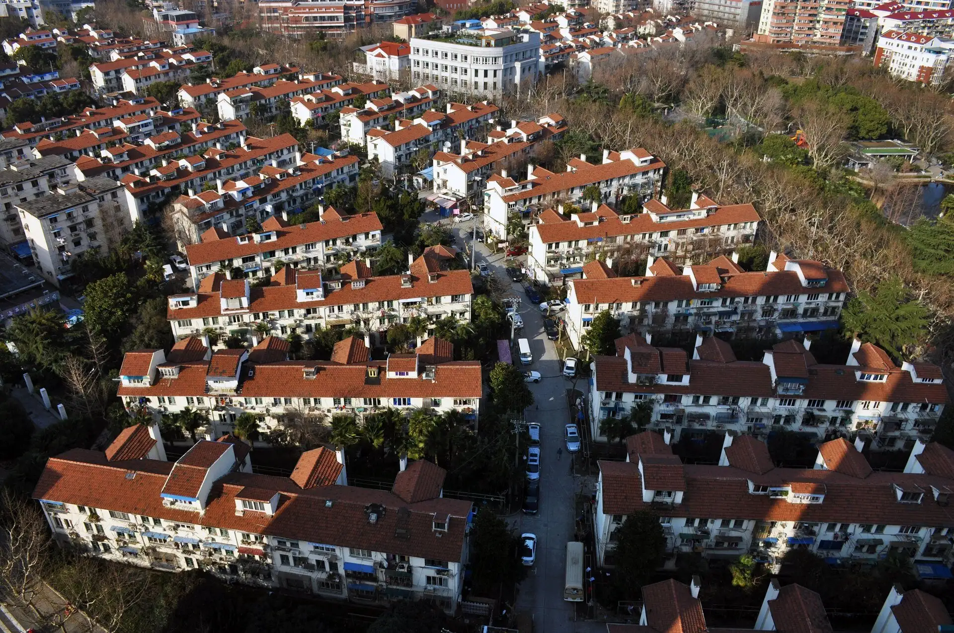 Aerial view of rows of white three-storey houses with red tiled roofs and dormers along a narrow lane lined with parked cars, trees and a park to the right.