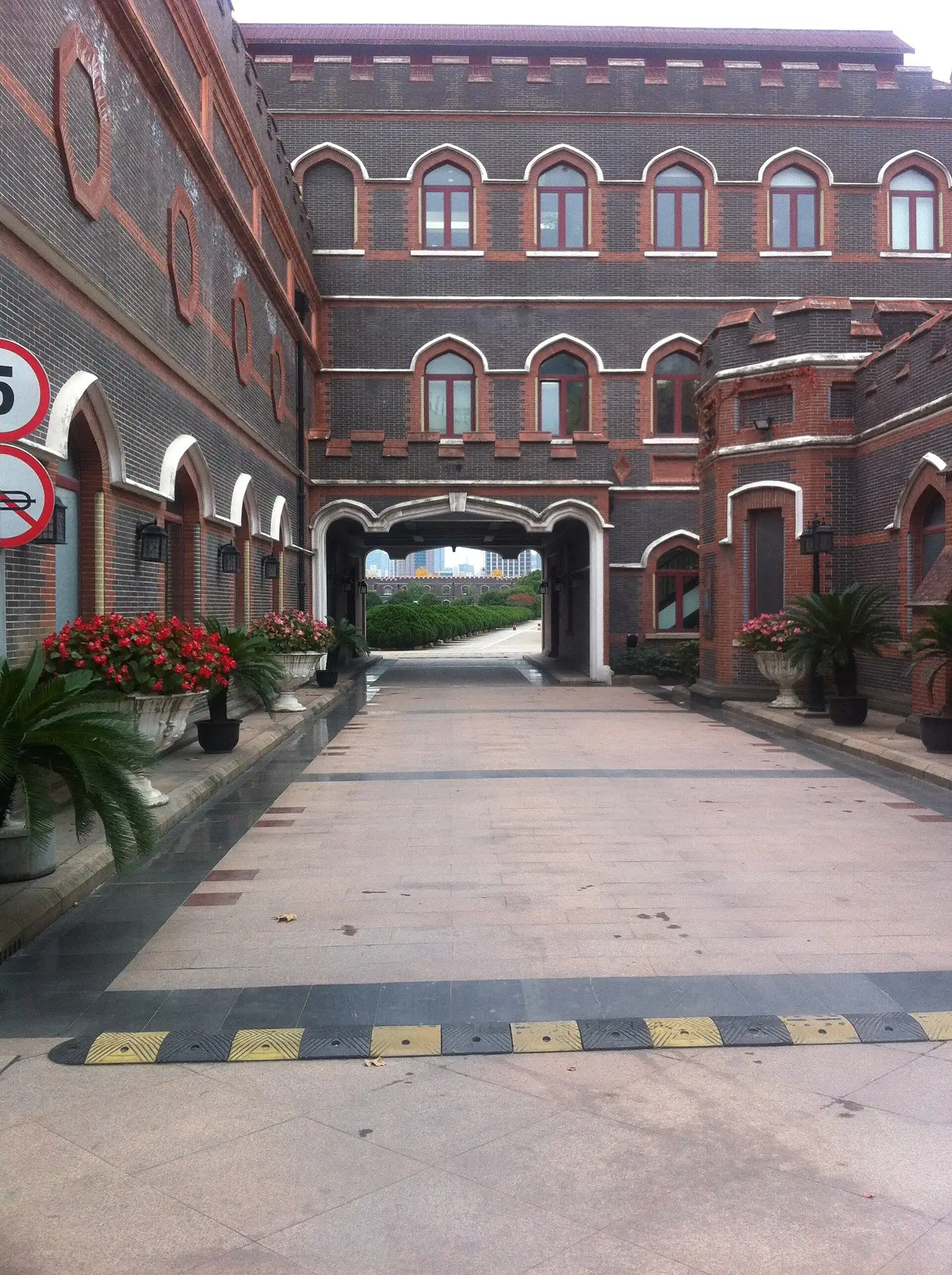 Paved courtyard between dark brick buildings trimmed in red brick, with pointed arched windows, crenellated parapets and an arched gateway leading to a tree-lined drive.