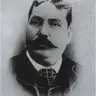 Black-and-white oval portrait of a heavy-set man with a dark moustache, wearing a checked tie and dark jacket.