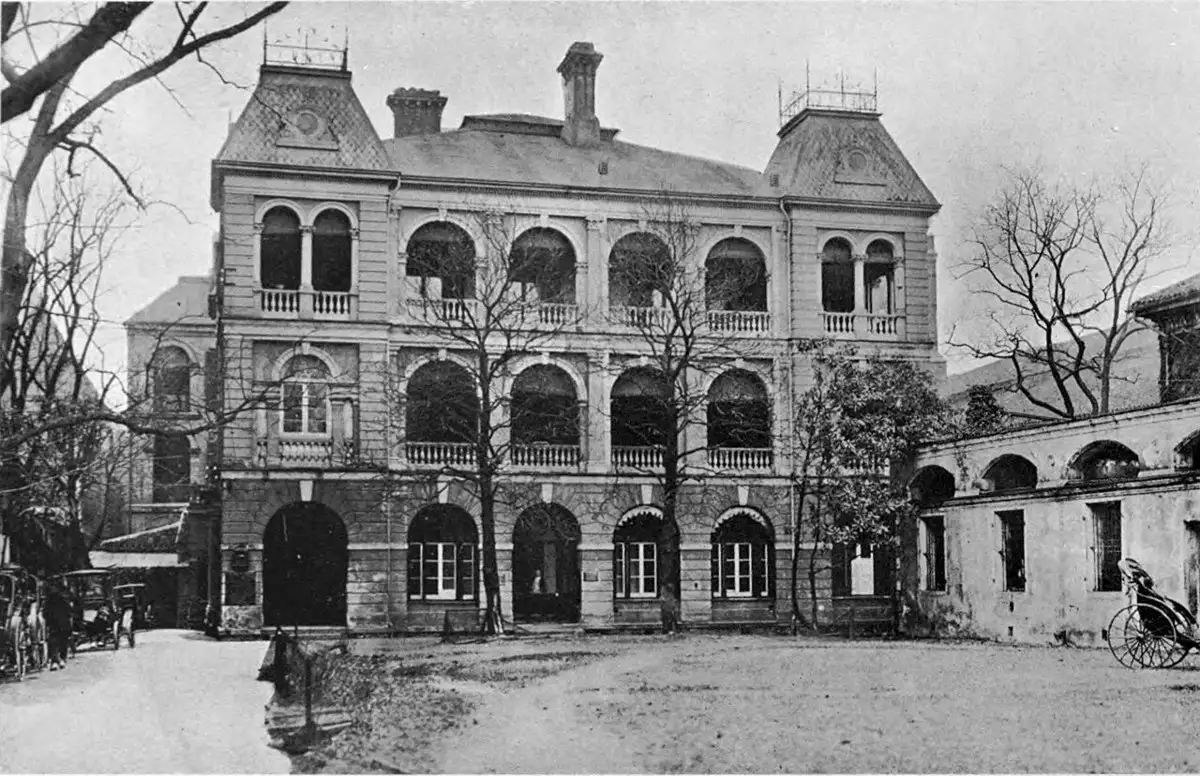 Black-and-white photograph of a three-storey brick-and-stone building with arched verandas, seen across a gravel courtyard.