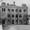 Black-and-white photograph of a three-storey brick-and-stone building with arched verandas, seen across a gravel courtyard.