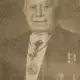 Faded newspaper-style photograph of an elderly man with a white moustache in evening dress with a sash and medals.