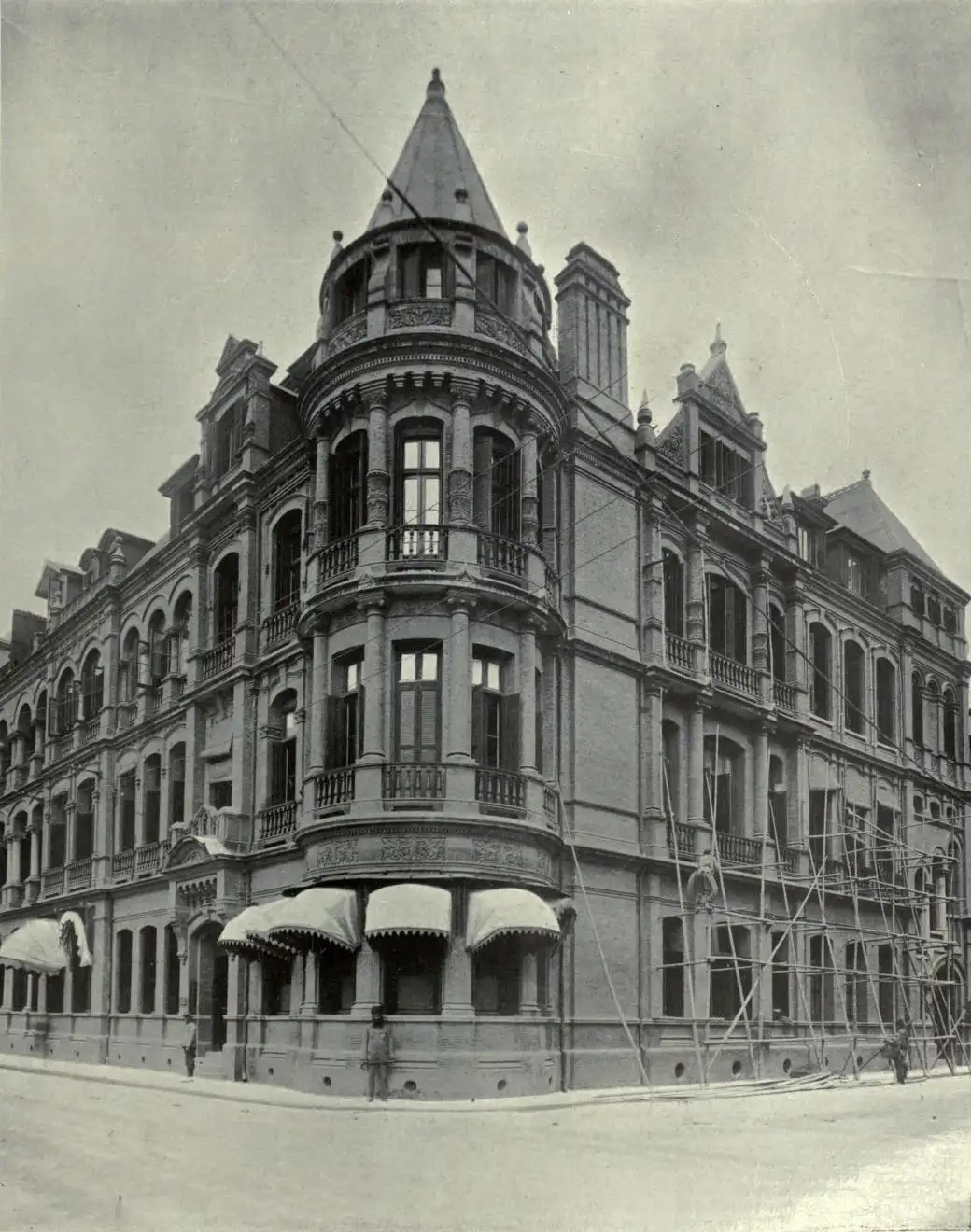 Black-and-white photograph of a corner building of red brick and stone with a conical tower, balconies and bamboo scaffolding on one side.