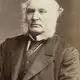 Sepia studio portrait of an elderly man with white mutton-chop whiskers, wearing a dark coat.
