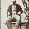 Sepia photograph of a bearded man in a dark jacket and bow tie, seated on a chair beside a potted plant.