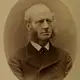 Sepia oval studio portrait of a middle-aged man with a bald head and long curly side-whiskers, in a dark coat.