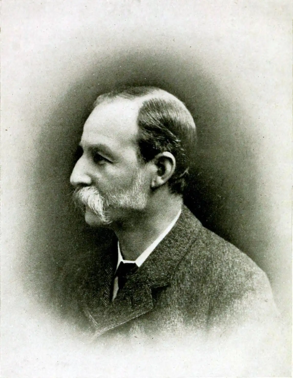 Black-and-white profile portrait of an older man with a white moustache and short beard, facing left, in a tweed jacket.