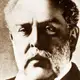 Sepia close portrait of a man with swept hair and a grey beard, in a dark coat.