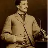 Sepia seated studio portrait of a young man with a moustache in a light suit, looking to the right.