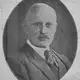 Oval black-and-white portrait of a balding man with a moustache in a suit and tie.
