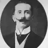 Black-and-white oval portrait of a man with an upturned moustache, in a dark jacket and high white collar.