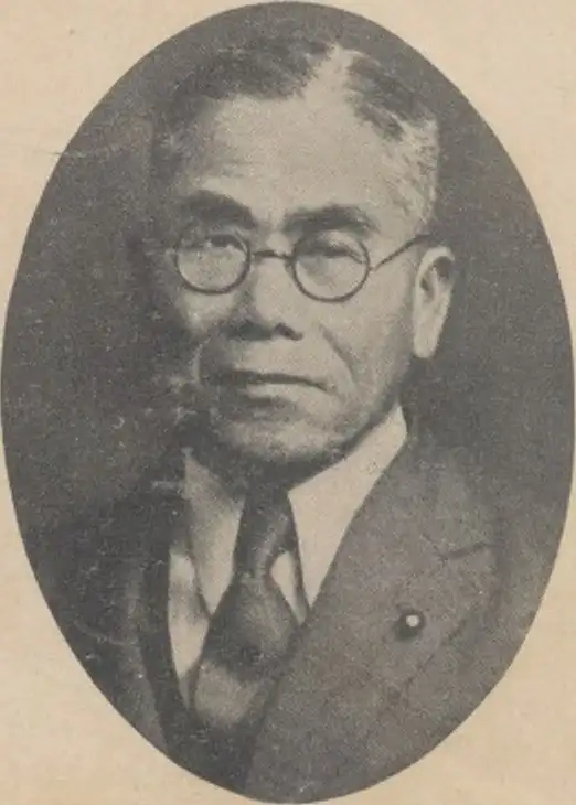 Black-and-white oval portrait of an elderly man in round glasses, jacket and tie, with a small pin on the lapel.