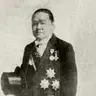 Full-length black-and-white studio portrait of a man in a tailcoat holding a top hat, wearing several decorations, with a printed caption below.
