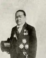 Full-length black-and-white studio portrait of a man in a tailcoat holding a top hat, wearing several decorations, with a printed caption below.