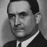 Black-and-white portrait of a dark-haired man in a dark suit and tie looking toward the camera.