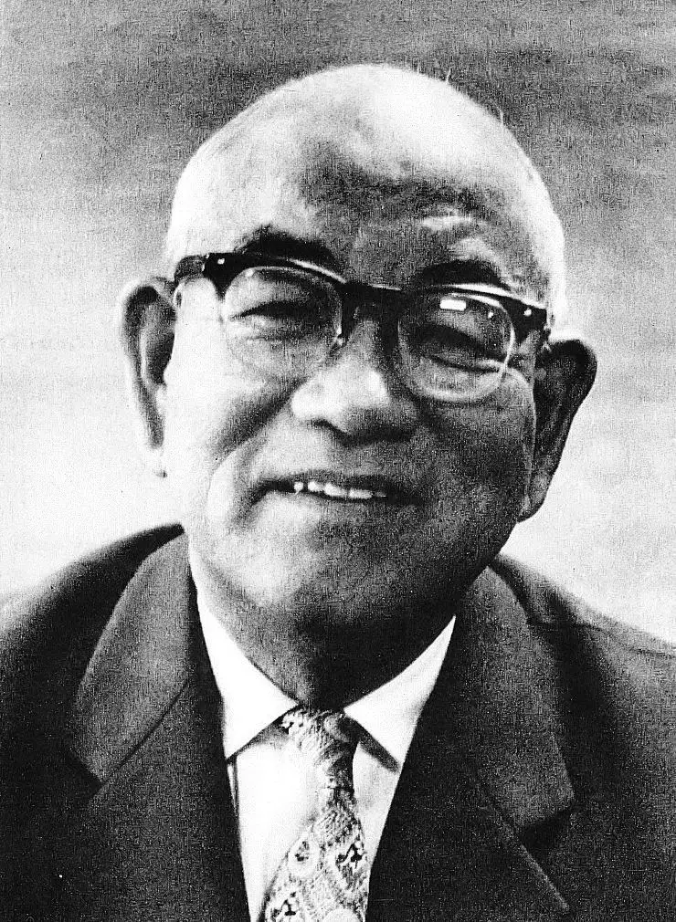 Black-and-white close portrait of a smiling bald elderly man in thick-rimmed glasses, jacket and patterned tie.