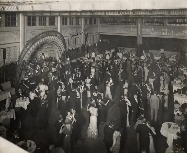 Old black-and-white photograph looking down on a large ballroom, couples in evening gowns and dinner jackets dancing, tables with white cloths round the edge, a band on a stage under a decorated arch at left.