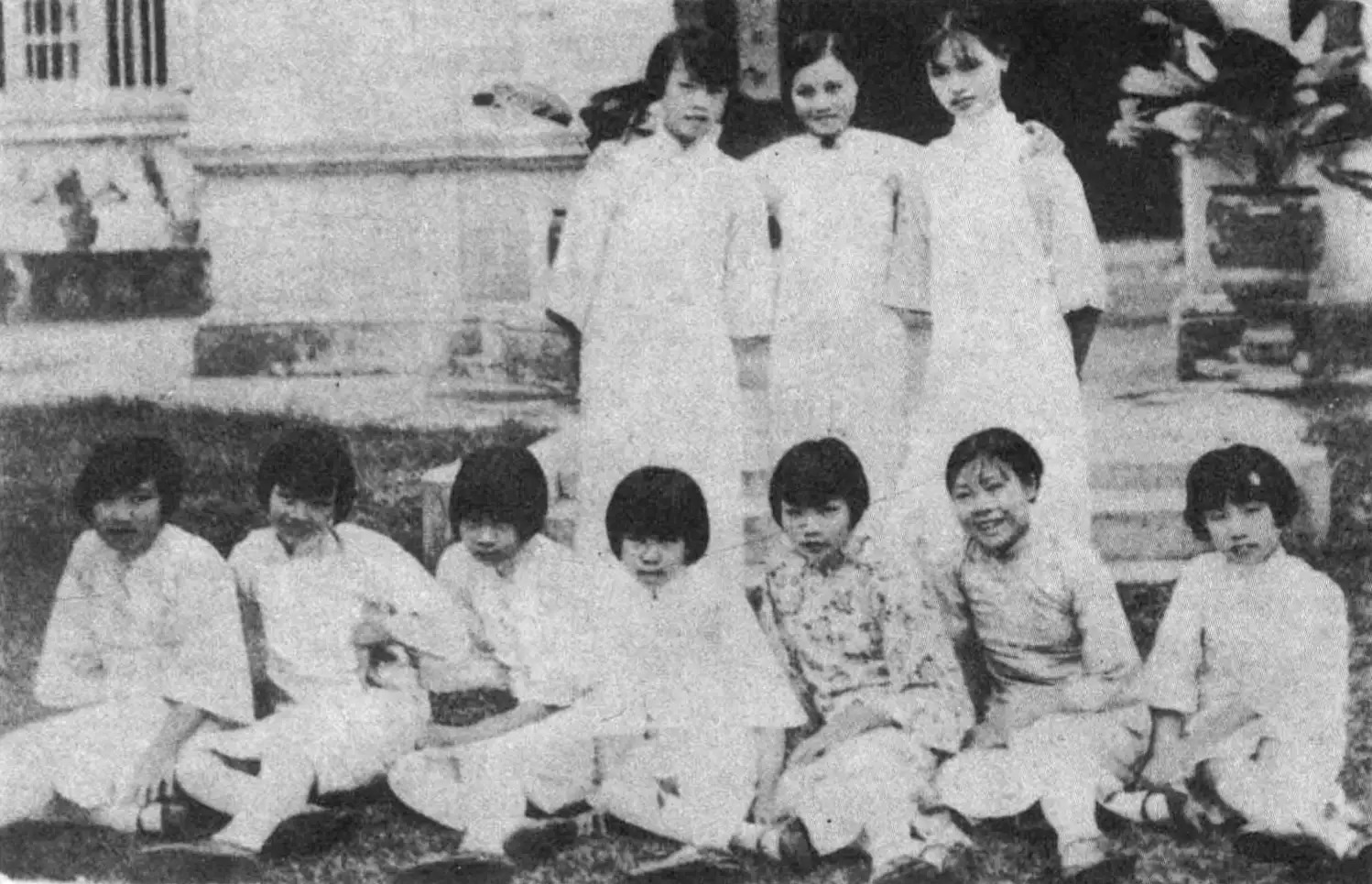 Black-and-white magazine photograph of ten young girls in white jackets and trousers, seven sitting cross-legged on a lawn and three standing behind them in front of a building.