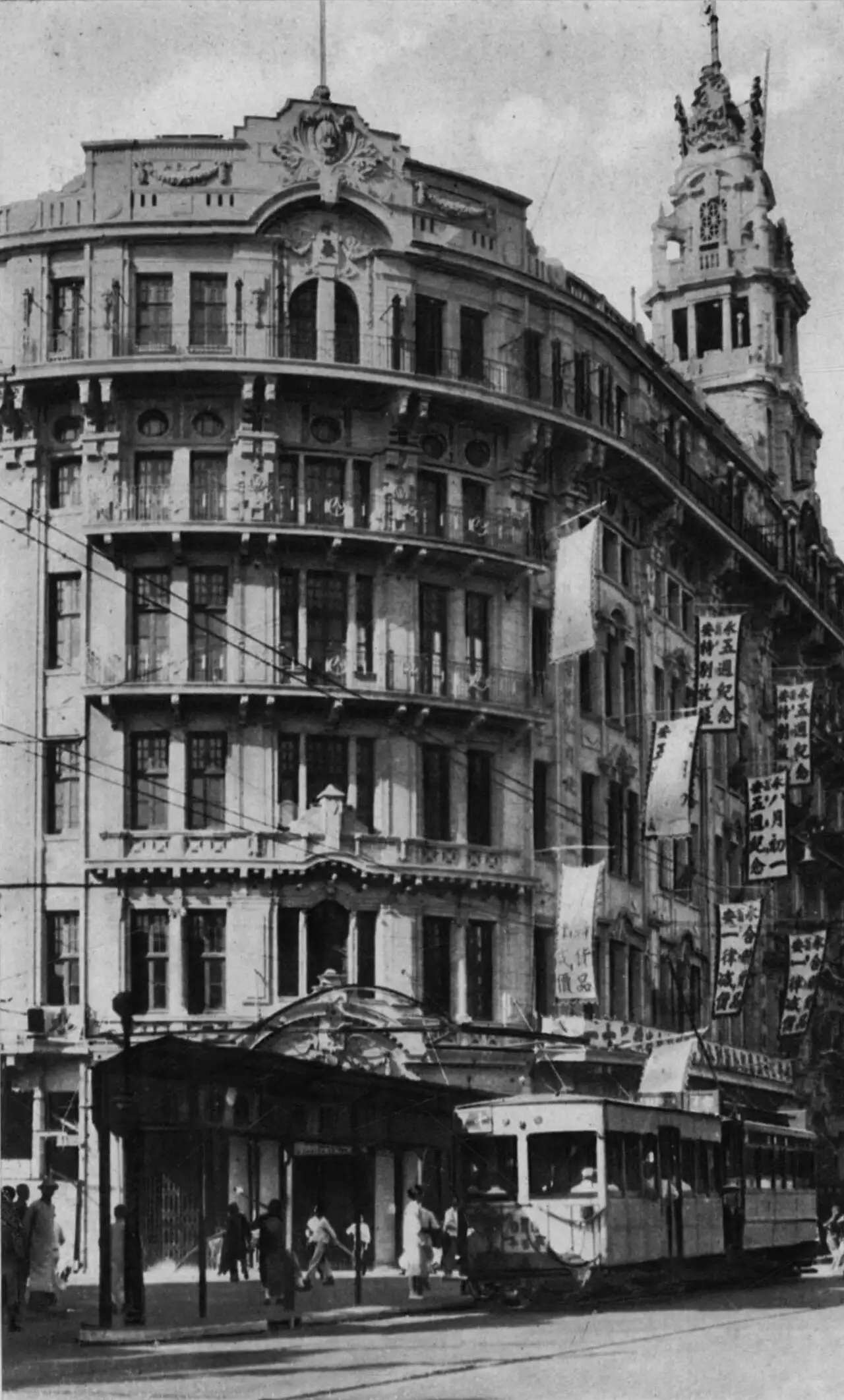 Black-and-white photograph of an ornate curved corner building with balconies on every floor and a tall decorated tower, vertical cloth banners with Chinese characters hanging down the front, and a tram in the street.