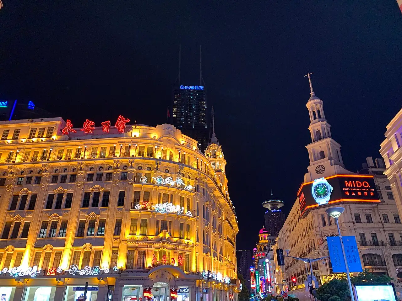 Night view of a floodlit ornate curved building with large red Chinese characters on the roof, a white clock tower on the right with an illuminated watch advertisement, a skyscraper behind.
