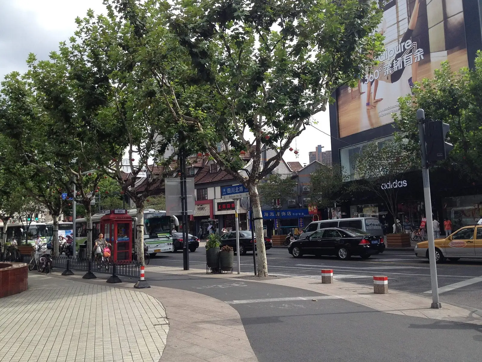 A tree-lined section of North Sichuan Road, with shops and pedestrians along the pavement.