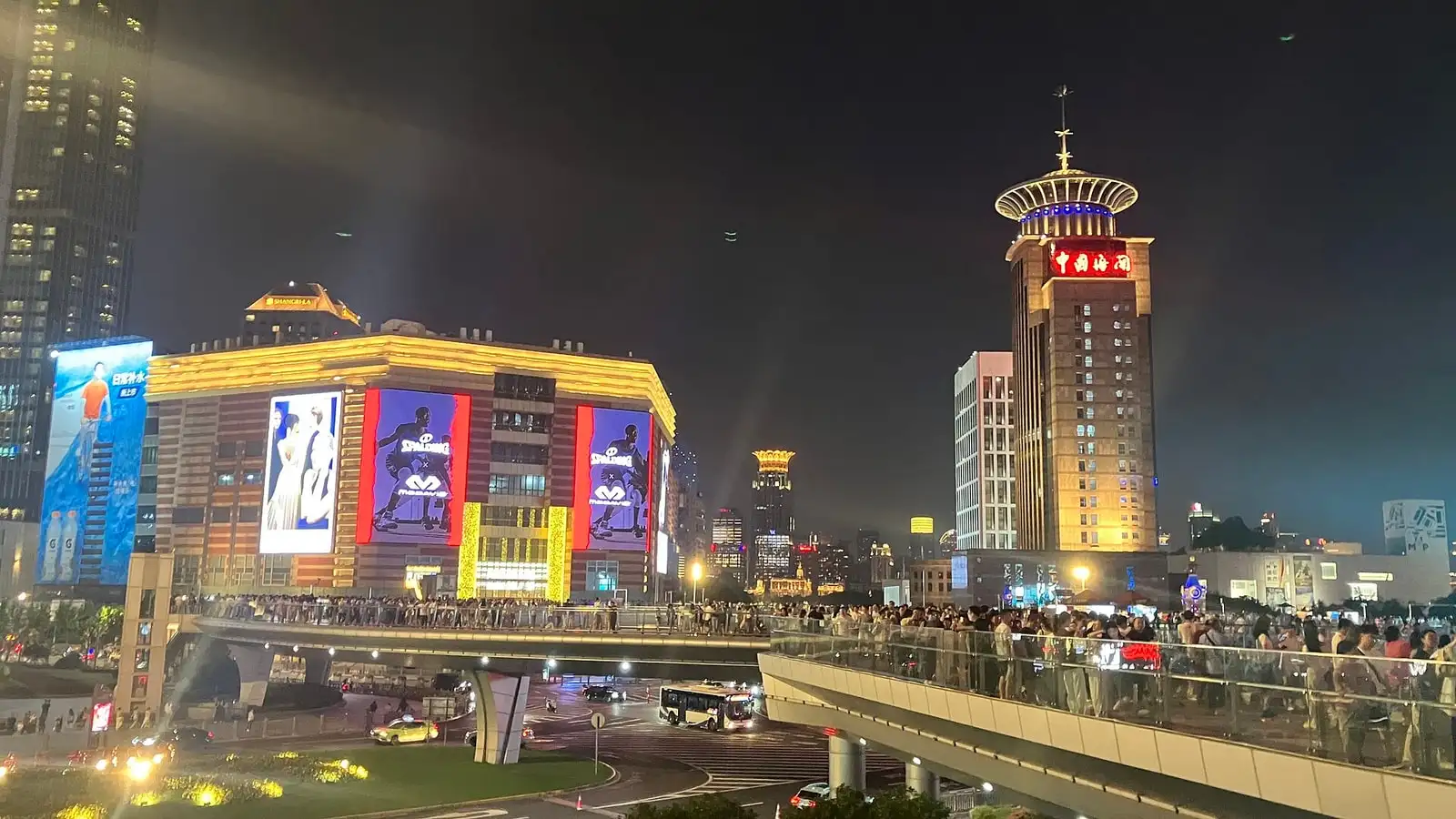 Super Brand Mall illuminated at night in Lujiazui, with a pedestrian bridge and the Oriental Pearl Tower behind.